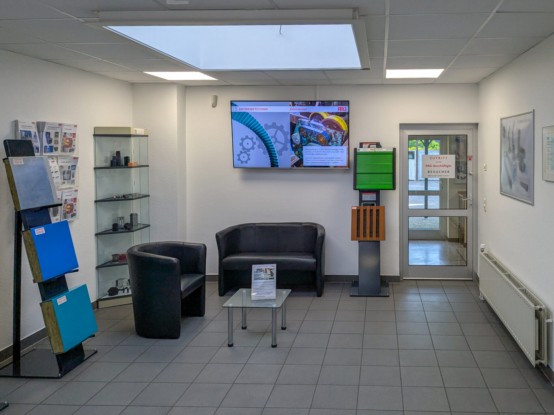 Empfangsbereich des RRG Ladenlokals mit schwarzen Sesseln, Sofa, Glastisch, Prospektständer, Glasvitrinen und Flachbildschirm mit Antriebstechnik-Präsentation