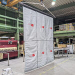 Sechs silbergraue Sonosorp Lärmschutz-Matten ECO als hängende Trennwand in einer Industriehalle, von oben an einer Traverse befestigt