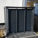 PREMIUM Sichtschutz aus anthrazit lackiertem Stahl mit Lamellenfront und seitlichen Lochblechen, installiert vor einer Außenwand auf Kies, zum Schutz und zur optischen Aufwertung einer Wärmepumpe.